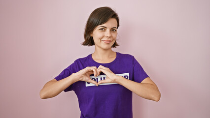 Smiling young hispanic woman volunteering, making love symbol with heart gesture, standing over isolated pink wall, confidently representing social support and community unity