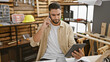 © Krakenimages.com - A focused man using a tablet and phone in a woodworking workshop, surrounded by tools and lumbers.