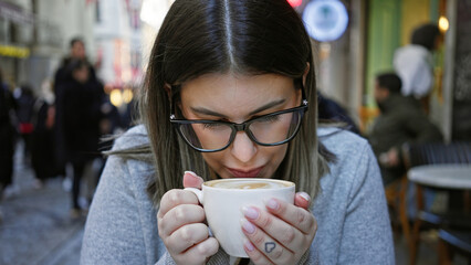 Wall Mural - A young woman wearing glasses enjoys a cup of coffee at an outdoor cafe terrace surrounded by urban activity.