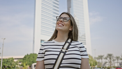 Canvas Print - Smiling young adult woman with glasses enjoying her time in downtown dubai among modern skyscrapers.