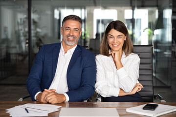 Wall Mural - Portrait of smiling at camera mature Latin businessman, European businesswoman sitting at table, workplace in office. Two diverse colleagues, team of confident man, woman professional business people.