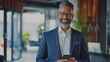 © chanidapa - Smiling happy confident mid aged male company ceo executive wearing suit holding cellphone standing in office using business mobile apps technology financial online solutions on cell phone.