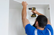 A man in uniform screws up an electric screwdriver to hang a kitchen cabinet on the wall.