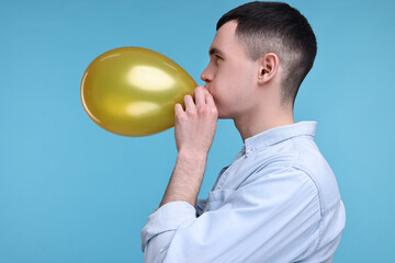 Wall Mural - Young man inflating golden balloon on light blue background