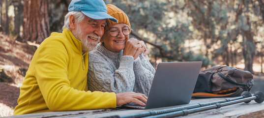 Head shot portrait close up of cute couple of old middle age people using computer pc outdoors sitting at a wooden table in the forest of mountain in nature with trees around them.