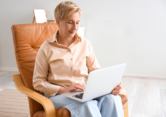 Mature female programmer working with laptop in armchair at home