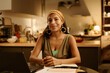 © pressmaster - Happy young Latino female freelancer or student looking at camera while sitting by kitchen table in front of laptop and networking