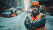 Construction worker in high visibility and reflective gear, safety vest and helmet stands with crossed arms on a snowy urban street, with blurred traffic and warning signs in the background.
