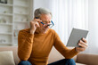 © Prostock-studio - Senior man adjusting his eyeglasses while using tablet at home