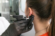 A doctor in black medical gloves pierces the ears of a beautiful young woman in the medical office using a safe black piercing gun and medical earrings