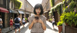 Asian young smiling woman, smartphone in hands, on cozy city street on sunny summer day, Film grain effect