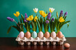Easter table centerpiece of colorful dyed eggs and yellow tulips and violet hyacinth Spring flowers in a cream ceramic egg carton on green background, on a dark wood table, front view.