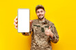 Ukrainian army soldier in military camouflage uniform shows smartphone screen and like on yellow isolated background, Ukrainian military cadet advertises phone and recommends