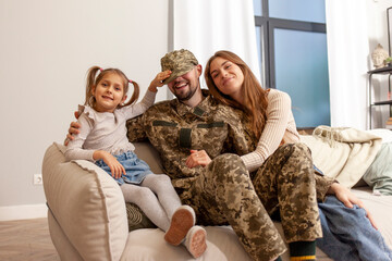 Wall Mural - Ukrainian military man in camouflage uniform hugs his family at home and smiles, a happy soldier of the Ukrainian army returned home and sits on the sofa with his wife and daughter, mobilization