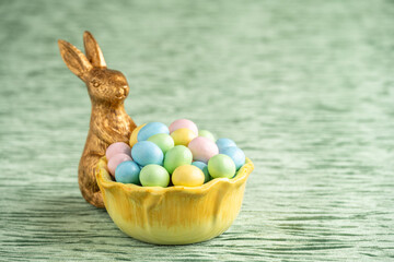 Wall Mural - Decorative gold Easter Bunny holding a yellow flower shaped bowl filled with candy coated pastel easter eggs on a textured green velvet background, Happy Easter
