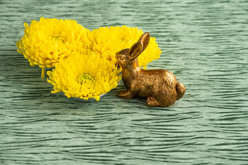 Wall Mural - Decorative gold Easter Bunny sniffing three large yellow chrysanthemum flowers on a textured green velvet background, Happy Spring
