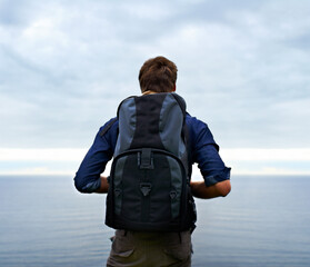 Wall Mural - Man, backpack and hiking by ocean for travel, sightseeing or outdoor journey in nature. Rear view of male person, hiker or tourist with bag on back for trekking, fitness or adventure with cloudy sky