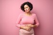 
Photography a middle-aged woman holding her belly with a worried expression, against a solid pastel pink background, portraying the discomfort of her stomach pain