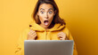 Surprised young woman in yellow hoodie reacting to content on her laptop