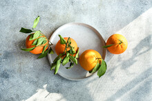 Top View Of Trio Of Ripe Oranges Adorned With Lush Green Leaves, Presented On A Grey Circular Stone Plate Against A Textured Background