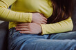 © Yura Yarema - Close up of unknown young woman suffering from menstrual pain. Girl with hands squeezing belly having painful stomach ache or period cramps sitting on sofa. Abdominal pain and painful periods concept