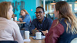 Casual Office Discussion Amidst Diverse Coworkers, Indoor Setting with Blue Wall Background, Informal Business Meeting with Four Individuals, Use of Technology, Coffee Break, Sharing Ideas and Casual 
