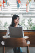 © Mongta Studio - Freelance concept, A smiling woman is working on her laptop in a cafe decorated with colorful paper lanterns, enjoying a bright day.