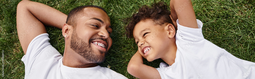 top view of cheerful african american father and son lying on green ...