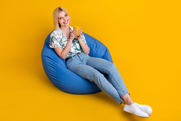 Poster - Full size photo of satisfied lovely girl wear jeans blouse sit on bean bag hold cup of cappuccino isolated on yellow color background