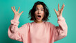 © Studio Nova - A woman with a surprised expression raises her hands in a dramatic gesture, wearing a pink sweater against an aqua background.