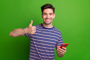 Wall Mural - Photo of handsome positive man toothy smile hold smart phone demonstrate thumb up isolated on green color background
