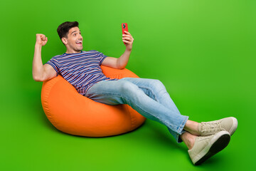 portrait of ecstatic man wear stylish t-shirt jeans sit on bean bag look at smartphone win lottery i