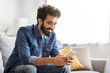 © Prostock-studio - Cheerful bearded indian man looking at his smartphone with smile at home