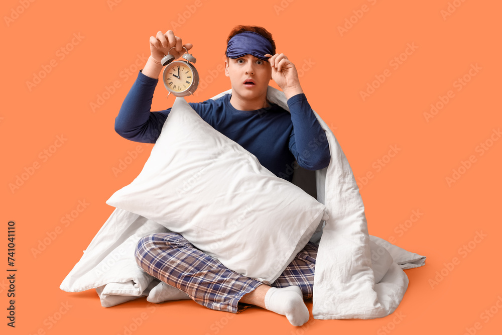 Handsome young shocked man with bedding, alarm clock and sleeping mask sitting on orange background