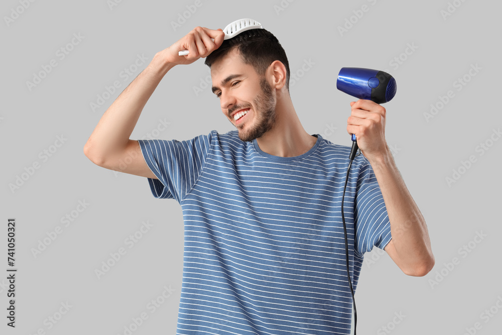 Handsome young man with hair dryer and brush on grey background