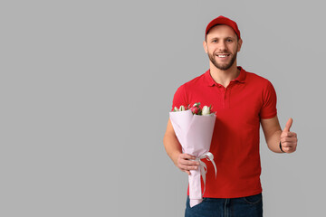 Canvas Print - Young delivery man with bouquet of beautiful tulips showing thumb-up gesture on grey background