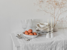 Retro White Ceramic Tableware, Glasses, Cutlery, Eggs In A Bowl On A Light Chest Of Drawers With A White Tablecloth