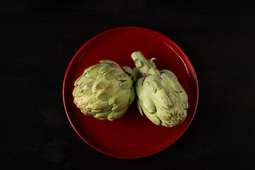 Aerial view of two artichokes on red plate, on dark wooden table, horizontal, with copy space