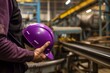 a factory operator holding a purple hard hat, with machinery and conveyor belt nearby