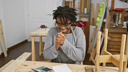 Wall Mural - Young, proud black woman with dreadlocks donning safety glasses at the carpentry workshop, jaw-droppingly ecstatic as she raises arm in joyful celebration of her exciting victory and success