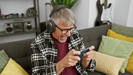 Canvas Print - Mature man in headphones plays with smartphone excitedly at home, expressing joy and excitement.