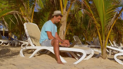 Freelance at beach. Rest and dreams at work, enjoying life while working at beach. Young man working on laptop computer on the tropical beach under the palm tree. Freelancer on sand in sun lounger.
