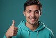 A young man in a turquoise hoodie gives a thumbs up, his friendly smile suggests approachability and positivity.