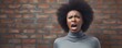 Frustrated African woman in gray sweatshirt against brick wall backdrop expressing anger. Concept Anger, Frustration, Portrait Photography, Brick Wall Background, African Women