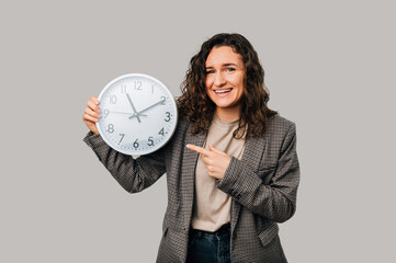 Cheerful young woman wearing jacket is holding and pointing at a round clock over grey background.