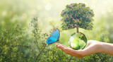 Fototapeta Kwiaty - Earth Day or World Environment Day, environmentally friendly concept. Tree growing on globe and Morpho butterfly in hand on green background. Save planet and protect nature, sustainable lifestyle.