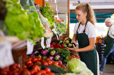 Young girl seller in uniform working in supermarket at her first job, checking organic eggplants