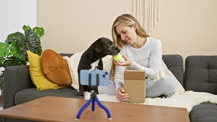 Wall Mural - A young blonde woman at home plays with her black dog, recording a video indoors with a smartphone on a tripod.