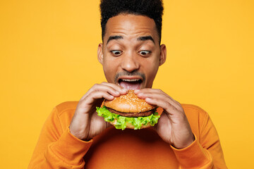 Wall Mural - Close up young surprised man wear orange sweatshirt casual clothes open mouth eat bite burger isolated on plain yellow background studio. Proper nutrition healthy fast food unhealthy choice concept.