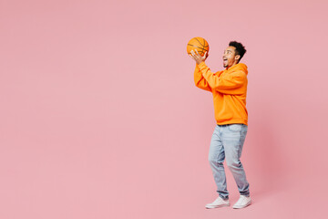 Wall Mural - Full body side view young man of African American ethnicity fan in yellow hoody casual clothes cheer up support basketball sport team hold ball watch tv live stream isolated on plain pink background